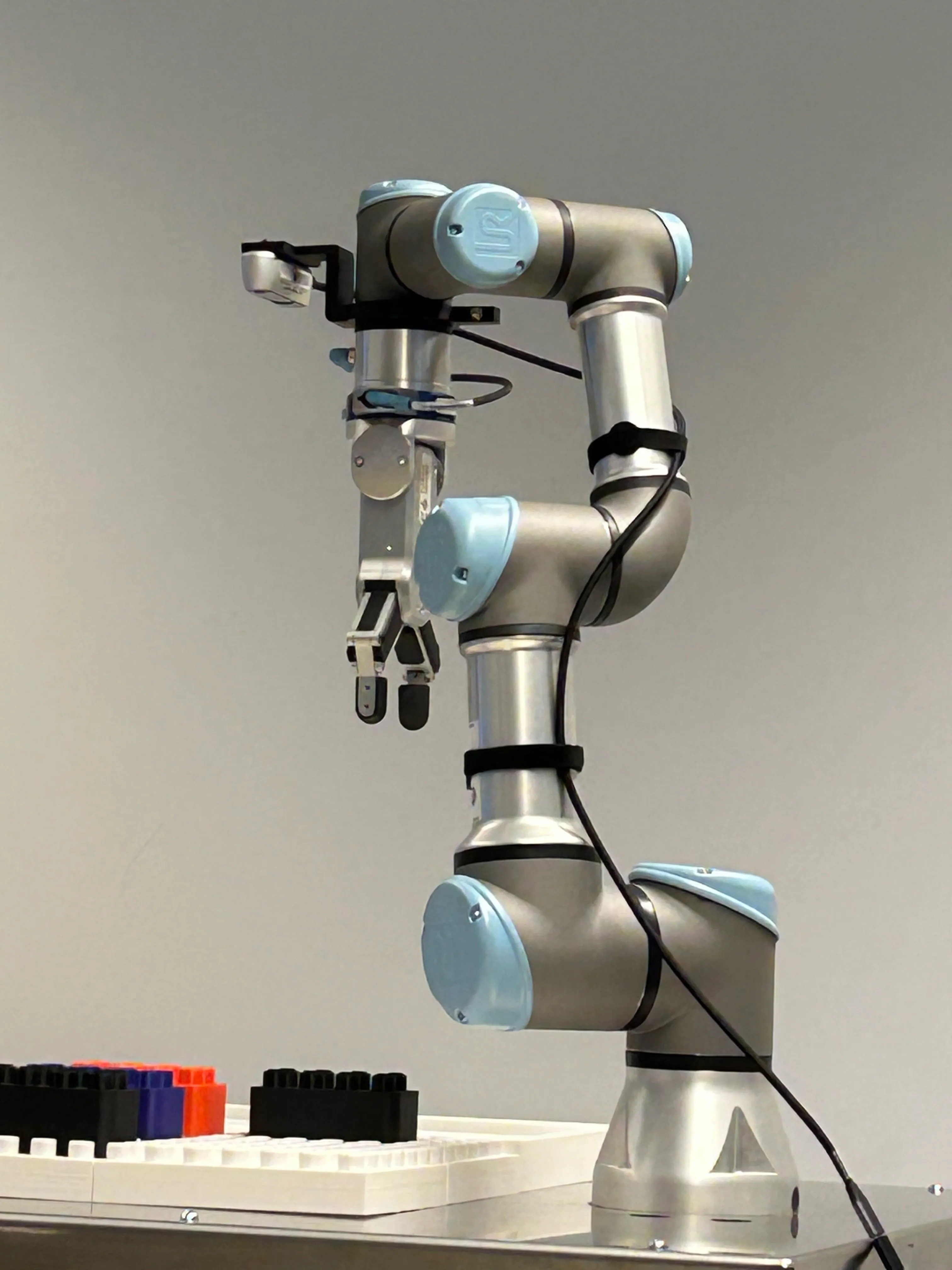 UR3e arm with a RealSense depth camera mounted above the wrist and a parallel jaw gripper below it, standing over a white base plate with black, blue and red bricks laid out beside it
